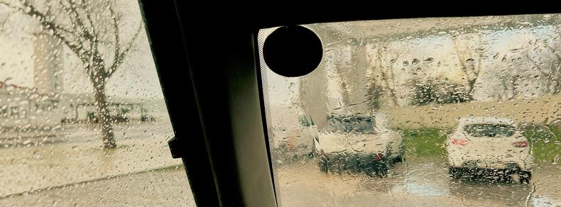 View through a wet car window with raindrops on the glass. The scene outside shows a street with parked cars and trees, slightly blurred by the rain. A tree and two white cars are visible. The image conveys a rainy day atmosphere.