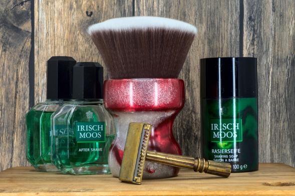 (Le Chat Mistral description)

This image features a collection of men's grooming items arranged on a wooden surface. There's a green bottle of "Irisch Moos" aftershave, a black tube of "Irisch Moos" shaving soap, and a gold-coloured safety razor. In the centre, there's a large shaving brush with a red and silver handle. The background is rustic wood, giving a classic, masculine feel to the setup. The items are neatly displayed, suggesting a focus on traditional shaving techniques.