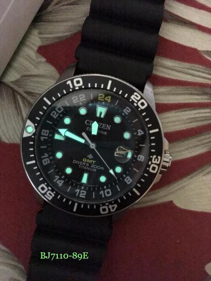 Somewhat noisy low light photo of Citizen BJ7110-89E (cal B877, c 2019) dive watch with matte black dial & luminescent main hands & hour indices which are glowing in light-mint color.

Second-time-zone hand, with yellow hollow arrow at the end, has no luminescence. It can only be adjusted in full 1-hour increments.

A 24-hour chapter ring is angled at ~85-65 degrees🤷🏽‍♂️. Hours with Arabic numerals are marked every 2-hour interval from "24" to "22"; in between are thin stick indices. Markers, except the "24" hour marker which is in light yellow, are in white.

The watch with a black rubber strap rests on a red/beige flower motif seat cover.

Time is 23:47:22.

Date window has been obscured by the second-time-zone hand; only some portions of current & next date digits are visible.

Tens place digit of the current date is 2; ones place digit could be 8 or 9. (Based on last modified date of the photo, current date is 28 that is about to turn over.)

Time in second time zone is 05:47:22.

The pip of the rotating bezel is at 9 o'clock position.

The watch is on Bonetto Cinturini 285 rubber strap (at the time of the photo).
