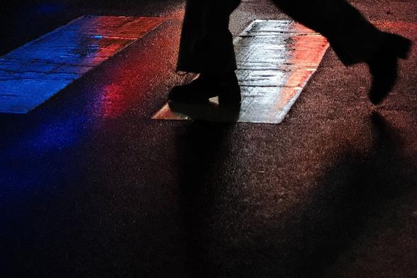 Eine Person überquert einen Zebrastreifen bei Dunkelheit. Nur die Beine und 2 Streifen sind zu sehen. Rotes, blaues und weißes Licht wird vom feuchten Asphalt und den Streifen reflektiert. Der vordere Fuß berührt den Boden, der hintere ist in der Luft und durch die Bewegung unscharf abgebildet. Die Beine sind nur als Silhouette zu sehen. 

A person walks over a zebra crossing in the dark. Only the legs and 2 stripes are visible. Red, blue and white light is reflected by the damp asphalt and the stripes. The front foot touches the ground, the rear foot is in the air and blurred by the movement. The legs can only be seen as a silhouette.
