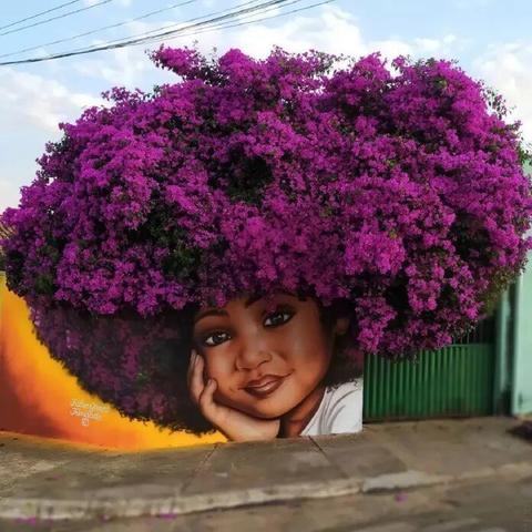 A stunning mural by Brazilian street artist Fábio Gomes Trindade in Rua Seis, Conjunto Arco Iris, Trindade, Goiás, Brazil. The artwork masterfully integrates a painted portrait of a young Black girl with the natural foliage of a tree above, creating the illusion of her curly afro hair. Trindade’s signature style blends street art with nature, turning everyday landscapes into powerful and poetic compositions. The vibrant colors and delicate details in her facial expression add depth and emotion, making this piece a striking example of urban art seamlessly coexisting with the environment.
