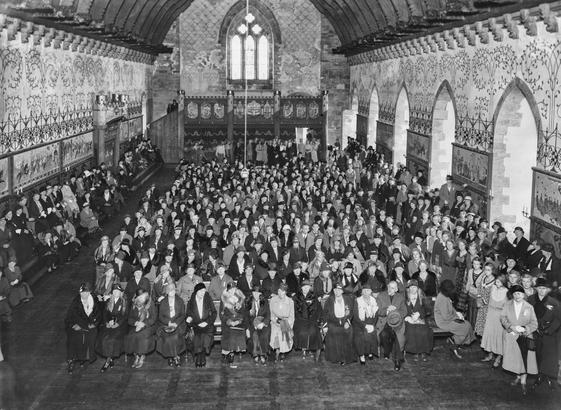A black and white photo of a large medieval stone hall filled with chairs of people, mostly women.