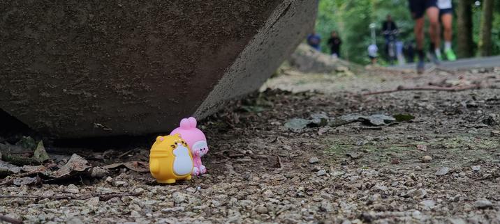 A photo of two tiny toys, an orange cat and a pink bunny made of silicone or rubber. They are sitting on some gravel next to some leaves at the base of a large trash can watching people job and bike by. They are about the size of a lego mini figurine. You can see lots of green trees and ivy in the distant background.