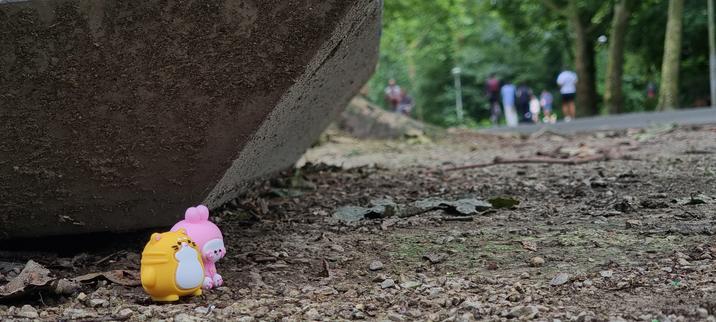 A second photo of two tiny toys, an orange cat and a pink bunny made of silicone or rubber. They are sitting on some gravel next to some leaves at the base of a large trash can watching people job and bike by. They are about the size of a lego mini figurine. You can see more of the green trees and ivy in the distant background.