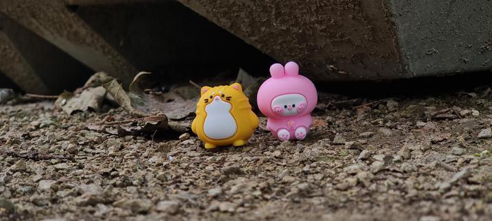 A third photo of two tiny toys, an orange cat and a pink bunny made of silicone or rubber. This one is a close up of the toys from their fronts. The orange cat has a mischievous expression and the pink bunny looks happy and excited. You can see the pink bunny's little toy beans. They are sitting on some gravel next to some leaves at the base of a large