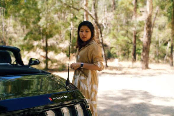 A woman stands in the middle of the frame, turning towards us and looking towards the rear of a car that’s to the left of the frame, with the right-hand tail lights in the middle of the bottom of the frame. Some trees are in the background, and it feels a bit dusty.
