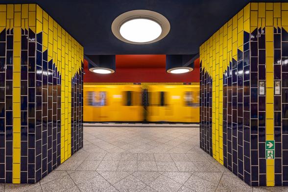 Symmetrical shot at slow shutter speed of a yellow metro entering the Richard Wagner Platz U-Bahn Station in Berlin, as seen between two walls separating the platform. The wall on each side of the picture frame are identically covered with ceramic tiles picturing a geometrical yellow palm tree on a dark blue background. Only two half wagons are visible between the walls displaying a quasi perfect symmetry. On the ceiling A big round lamp appears in the middle on a dark blue background in the foreground and and two other lamps are hanging in the background above the wagons on a red ceiling, completing the symmetry.