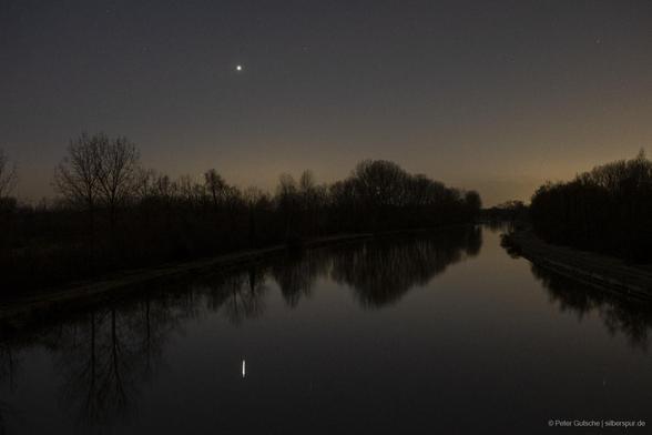 A calm river reflecting the fading light of dusk after sunset. The water's surface is smooth, mirroring the deepening hues of the evening sky. Above the distant tree line on the riverbank, the bright planet Venus shines prominently, its reflection shimmering on the water. Near the yellowish horizon, just above the trees, the faint glow of Mercury is also visible, adding to the serene and celestial atmosphere of the scene.