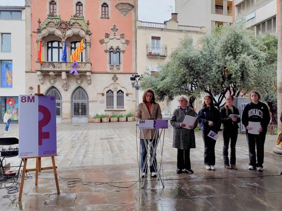 Cinc dones d'edats diferents, sota la porxada d'un dia lleugerament plujós, fan la lectura del manifest institucional del 8 de març al costat del cartell i just rere seu és veu la façana de l'ajuntament de Granollers amb el llaç lila penjat al balcó.