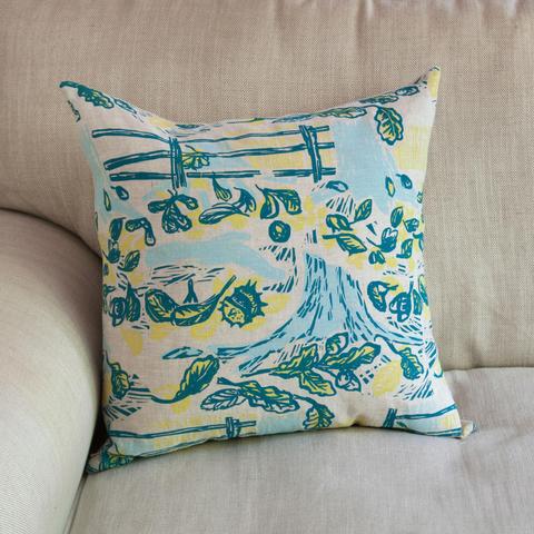 A hand printed cushion with fence, conkers, leaves and tree stump printed on it in blue and yellow.