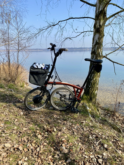 Ein Brompton steht an einem Baum. Im Hintergrund ist der Cossi zu sehen.
