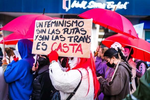 Fotografías de mujeres con sombrillas rojas en una manifestación a favor del trabajo sexual, con una mujer con máscara blanca mostrando una cartulina con la leyenda «un feminismo sin prostitutas ni transexuales es una casta». / Photographs of a women with red-umbrellas at a sex-work positive manifestation with one white mask wearing woman displaying a cardboard with "a feminism without hookers and transsexuals is caste" written over it.