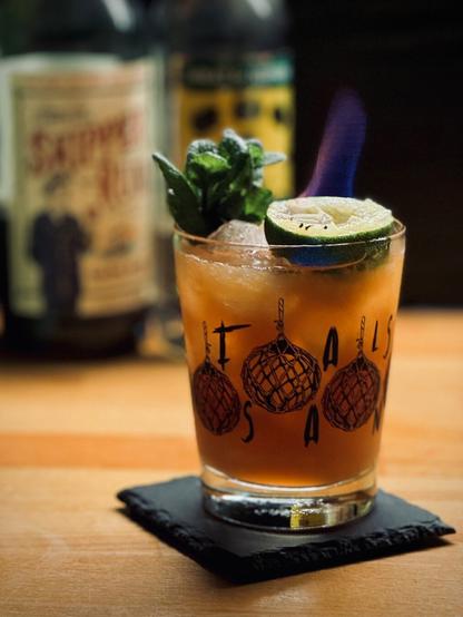 light brown cocktail with mint sprig and flaming spent lime shell garnish in a False Idol mai tai glass sitting on a black slate coaster with bottles of Skipper's Finest demerara rum and Wrap & Nephew overproof pot still rum in the background