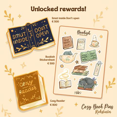 The image has a large title that reads "Unlocked rewards!" Below that we can see 2 golden enamel pins and a cozy book stickersheet. The first pin is a book laying open with on the back and front the text "Smut inside. Don't open." on a deep blue cover. The second pin is a book laying shut with on the front cover the words "Cozy reader" on a warm brown cover. In the bottom right corner we see the words "Cozy Book Pins Kickstarter”