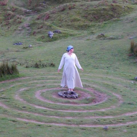🇩🇪 Das Foto zeigt eine weibliche Person, die inmitten einer Grasspirale steht. Sie trägt ein langes, helles Kleid und ein blaues Kopftuch und ist barfuß. Die Spirale befindet sich auf einer Wiese, im Hintergrund ist ein grüner Hügel zu sehen. 🇬🇧 The photo shows a female person standing in the middle of a grass spiral. She is wearing a long, light dress and a blue headscarf and is barefoot. The spiral is in a meadow, with a green hill in the background.