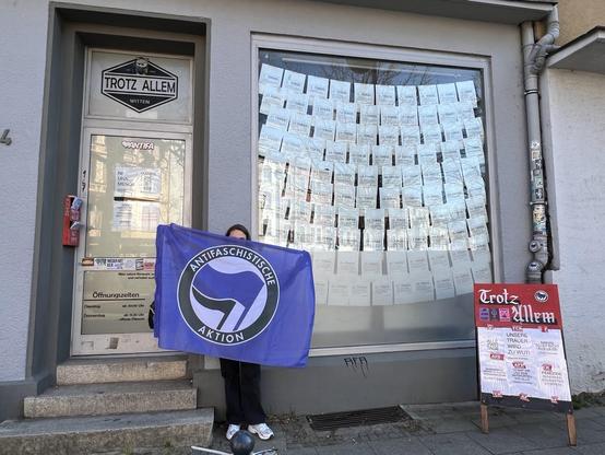 Tür (leider nicht barrierefrei) und Schaufenster des Trotz Allem: Im Schaufenster sind viele A4-Zettel mit Beschriftung aufgehängt. Eine Aufstelltafel mit Beschriftung „Trotz Allem“ steht rechts vom Schaufenster. Zwischen Tür und Schaufenster steht eine junge, weiblich gelesene Person mit einer Flagge der Antifaschistischen Aktion.