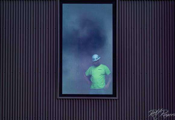 A man in yellow construction t-shirt and  white hardhat looks out window of a building being renovated.