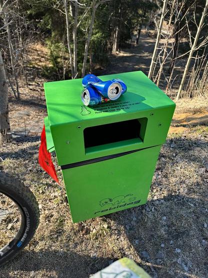 Auf einem giftgrünen Robidog (Abfalleimer für Hundescheisse verpackt in Plastiksäcke) liegen zwei leere Feldschlösschen Bierdosen die zuvor noch mitten im Wald lagen. Bald finden sie ihren Weg in den Abfalleimer.