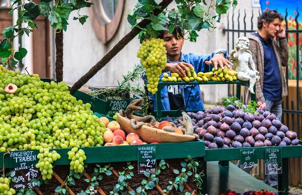 A market stall bearing fruit, such as grapes, bananas, figs, peaches and with green vine type leaves twining around the stall. There is a bowl with what seems to be two snakes, facing each other with mouths open. I think they are toys, probably. The vendor,, standing behind the stall is arranging small yellow bananas and at the end of that shelf is a white statuette of a cherub. There are price tickets displayed and behind the stall is a man talking on his mobile phone next to a black iron grating.