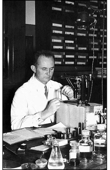 image/jpeg a historical photo of clean cut man in a lab coat sitting in front of a microscope in a lab. Photo from Max Planck Institute of Evolutionary Biology.