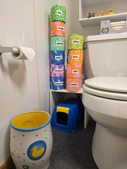 Picture of a bathroom toilet. In the center of the picture are ten stacked bamboo WhoGivesACrap toilet paper rolls, each with one word on the paper wrapper on the rolls spelling out: my magic planet with banana tentacles play love wet water. The rolls are sitting on a white shelf along with two stacked buckets. Under the white shelf is a blue stool with a yellow handle. R side: full left half of a white ceramic toilet with a closed lid and silver handle, a box of tissue is sitting on the back with a sticker on the box of two kissing fish. Above the tissue box is a white shelf with three items: a small plastic rubber duck with a snorkel, a Lego duck, and a MegaBloks Dispicable Me Jelly Lab including Minion Bob on a train hauling a batch of purple bottles. L side: on the floor is a white (outside) and yellow (inside) ceramic garbage can decorated with blue bubbles and a yellow duck. Above the garbage can is a toilet paper roll with a "triangle fold" fancy fold.