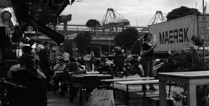 Cluttered view along a restaurant strip with motorbikes, trucks and wharf cranes in the background.