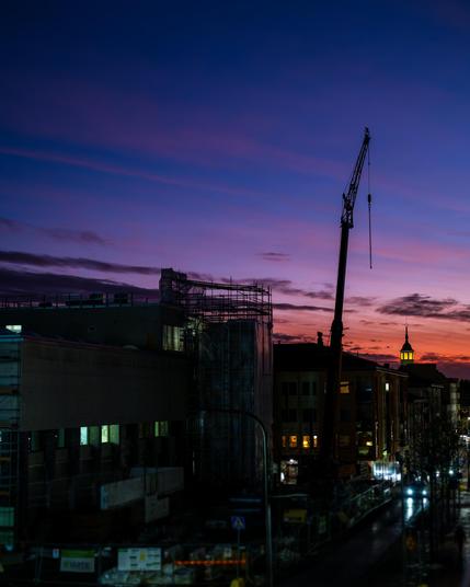 The image depicts an urban scene at dusk. There is a construction site with a crane prominently visible, surrounded by buildings. The sky has a gradient of colors from deep blue to orange, indicating sunset. Some lights are on in the buildings and along the street, suggesting evening time. The overall atmosphere is a mix of urban development and a calm evening setting.
