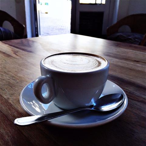 "This picture shows a cup of coffee on a table. The coffee appears frothy and has latte art on top. There is a spoon near the cup, suggesting that it might be a cold brew coffee. The setting suggests a café or coffee shop environment."