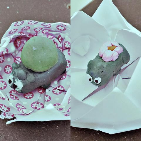 "This picture shows a creative handmade project featuring a small frog figurine adorned with a green leaf on its head, sitting on a flowery surface, placed within a cardboard box. The overall mood of the image is cheerful and whimsical."