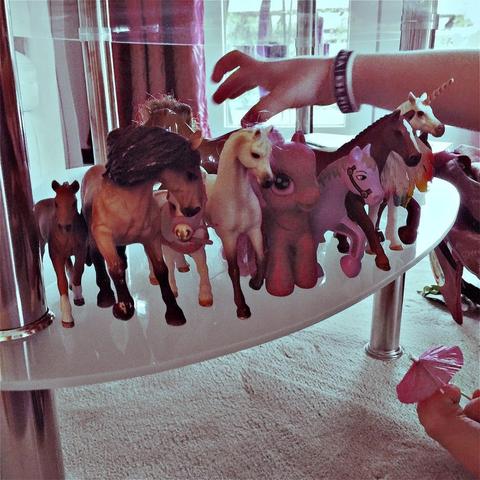 This picture shows a collection of colorful plastic horses of varying breeds, presented on a white surface. There is an action of reaching for one of the horses. The mood appears playful and child-friendly.