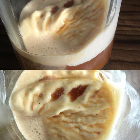 This picture shows a close-up of a glass cup filled with a beige-colored frothy liquid. The cup appears to be on a table, as indicated by the partial view of a tabletop at the bottom of the image. There is a visible spoon resting within the drink.