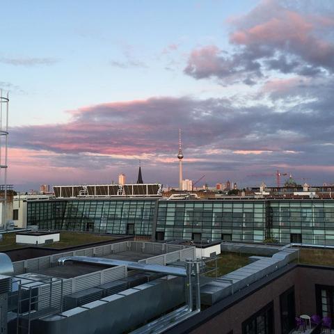 This picture shows a city skyline at sunset during early evening, as indicated by the soft pink hues. The sky is dotted with clouds, and there's an urban building complex in the foreground. In the distance, you can see silhouettes of buildings against a darker background that suggests it might be nighttime or near sunset. There are also a few visible structures, possibly cranes or construction equipment, hinting at ongoing development or urban expansion. The overall mood is serene and picturesque, capturing the beauty of an urban landscape as the day transitions to evening.