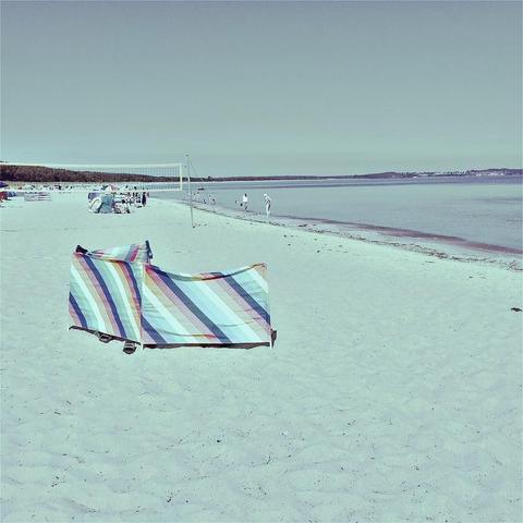 "This picture shows a beach scene with sandy shores, a striped beach towel laid out, umbrellas, and several people enjoying the day by the water. The mood is leisurely and sunny."