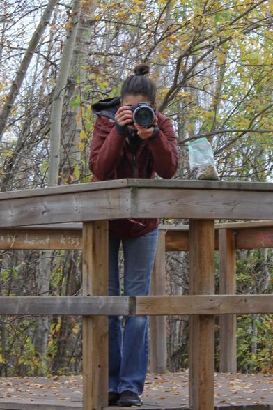 Photo of a woman taking a photo of the photographer. She is standing on a wooden lookout in a forested nature conservatory. Photo credit K.L.