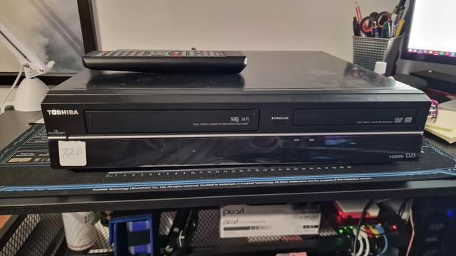 Photo of the front of a Toshiba DVR19DTKB2 combination DVD and VHS recorder, sitting on my desk. The unit is all black with a thin silver trim horizontally across the length of its front panel. The chassis is metal, and the front panel is plastic with a black glass pane across the entire bottom half, which still has its protective film on it. There's also a white sticker on the glass with hand-written number "720" on it. The remote control is sitting on top of the unit.