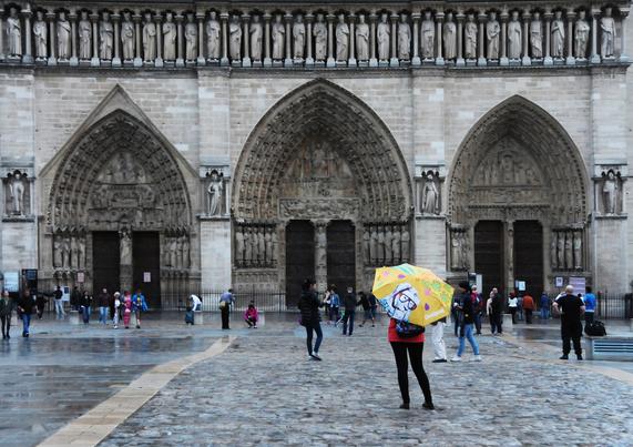 People walk in front of Notre Dame - there are three ornate doorways with draped figure statues inside the doorways and in a row above.on the right lower corner is a figure in a red jacket with a bright yellow umbrella. The umbrella has a face with a snorkel on it.