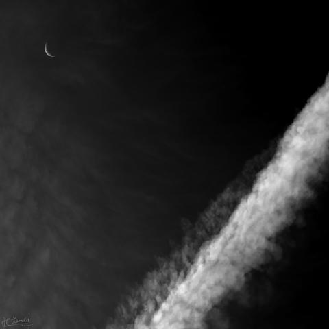 A square black and white image of the sky during the day, which is almost black in this processing. A few thin veil clouds can be seen on the left and the crescent moon on the top left. A broad, white contrail runs diagonally from the centre of the lower edge of the image to the centre of the right edge.