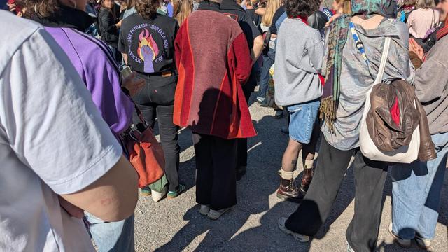 Crowd at a demonstration with different persons/colors/textures. Towards the left a print on a T-Shirt reads: Stop Femicides around a fist on flames.