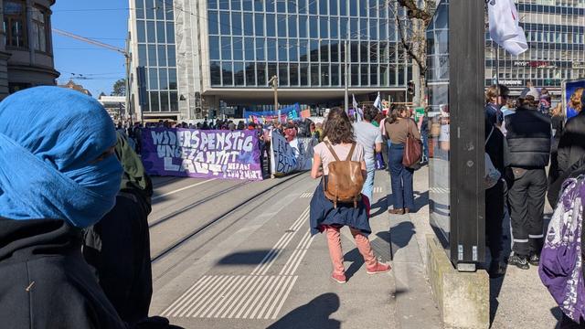 A Photo of a demonstration that is about to start. On the left a masked person with a blue cloth wrapped around their head. More poeple standing around. A tall building in the background in front of a blue sky. In the middle of the image a slogan written on a banner: Widerspenstig, ungezähmt, laut (english translation: Unruly, untamed, loud)