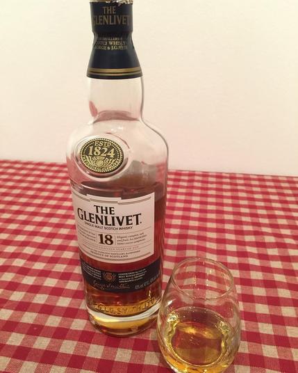 This picture shows a bottle of Glenlivet 18 Year Old Scotch whisky placed on a table next to a glass containing a small amount of amber-colored liquid, suggesting it is the whisky. The setting appears to be indoors with a plain wall and a checkered cloth visible in the background. The image likely conveys a sense of relaxation and enjoyment associated with savoring a fine whiskey.