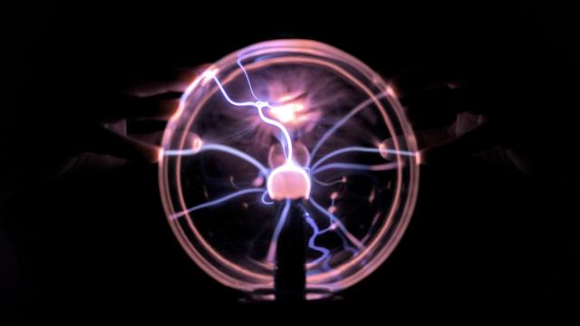 A photo of a plasma ball in darkness. There are fingers touching it and a face behind it.