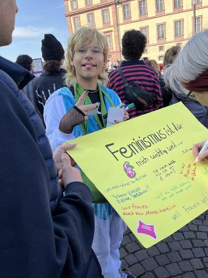 Eine Gruppe von Personen steht um ein hellgrünes Plakat mit der Überschrift "Feminismus ist für mich wichtig, weil ..." und eine Person schreibt mit rotem Stift etwas darauf.