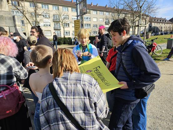 Eine Gruppe von Personen steht um ein hellgrünes Plakat mit der Überschrift "Feminismus ist für mich wichtig, weil ..." unterhalten sich.