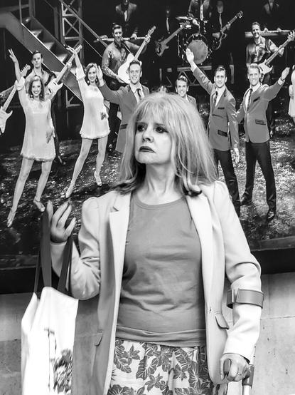 An elderly lady wearing a lot of make-up and carrying a crutch is standing on the pavement. Behind her is a poster for a musical that looks like it dates from the 60s, with smiling men and women.