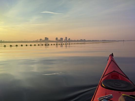 The front of a red kayak sits at rest on very calm water at sunset. The light is reflected in the still water, there are some tallish buildings on the horizon, and a line of round buoys bob in the foreground, the sky is yellowing in the setting sun with wispy clouds