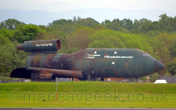 Photo of a green, but badly corroded, Fire Training Rig, a metal construction that vaguely resembles several different types of jet airliner, used for fire training, facing to the right.
The forward section looks very much like a Boeing 747-400, with an elongated upper deck, and a very short body section leads to an engine mounted on top of the fuselage, similar to a DC-10, but without the tail.
Shortened wings have an engine pod hanging underneath, and flaps appear to be extended to their full extent.
There are white "Simulation[dash]ters[dot]com" and "Manchester Airport" titles on the upper forward fuselage, the latter being rather faded, and "Fire Training Rig" on the tail engine.
Although mostly green, there are brown and orange rust marks all around the body, especially around the middle of the airframe and around the engine pods.
Green grass fills the foreground, with trees behind, under grey skies.