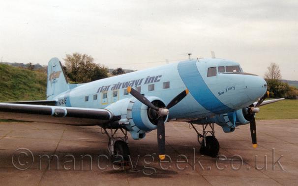 Side view of a light blue, twin propellor-engined World War 2-era passenger airliner parked facing to the right.
There is a darker blue band running around the forward fuselage, just aft of the cockpit, and another around the rear fuselage, just in front of the tail, as well as around the engine cowlings.
There are large black "Star Airways Inc" titles outline in gold on the upper fuselage, and a gold "Star" logo on the tail. 
The registration "N3455" is on the rear fuselage in black, and the name "Ian Drury" in a dark blue cursive script on the nose.
A grass bank behind the plane on the left slowly comes down to meet the ground on the right of the frame, with trees marking the airfield perimeter.
Hazy grey sky fills the rest of the frame.