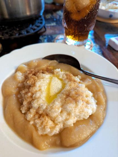Dark Table, white soup plate filled with rice pudding and a bit of butter and cinnamon and sugar on top. Around the rice pudding is apple Sauce. A spoon sticks into the apple sauce. Behind the dish is the cooking pot and a glas full of golden-brown liquid and ice cubes.