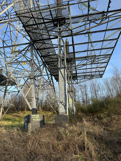 A photo of the base of an electricity pylon showing a connector that transports the power to a substation. A sign on the pylon says “Danger of death. Keep off”. There are bare trees in the background.