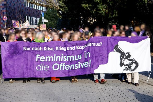 Viele Menschen hinter einem lila farbenen Transparent mit einer symbolisierten Frau* und mit dem Text "Nur noch Krise, Krieg und Kapitalismus? Feminismus in die Offensive!"